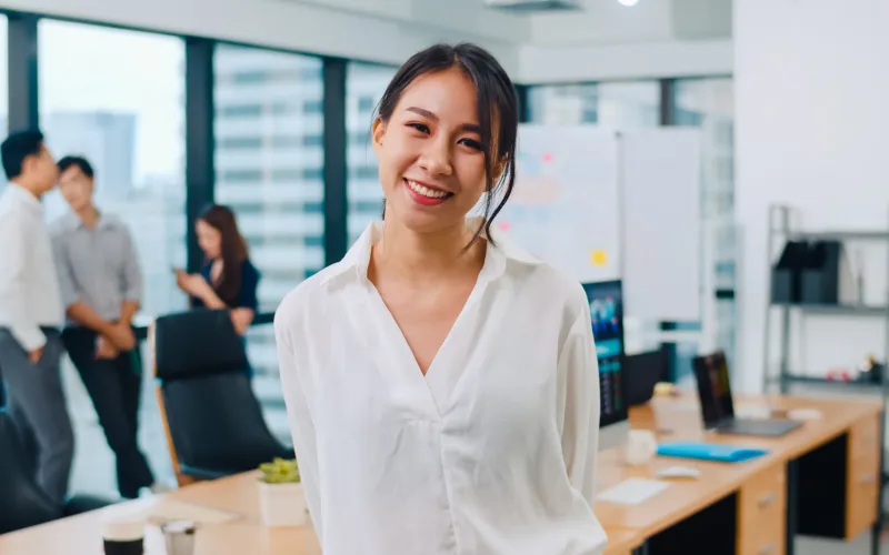Portrait of successful beautiful executive businesswoman smart casual wear looking at camera and smiling in modern office workplace. Young Asia lady standing in contemporary meeting room.